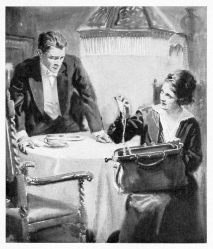 Man leaning over a table looking at a seated woman as she raises a necklace from a large purse in her lap