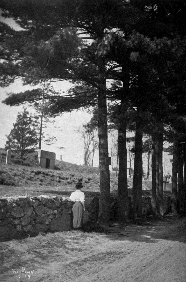 Helen is standing by a stone wall next to a dirt road