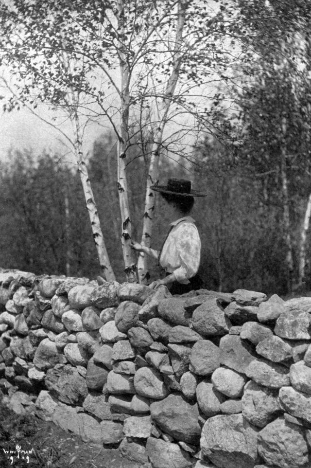 Helen wearing a hat is standing behind a stone wall