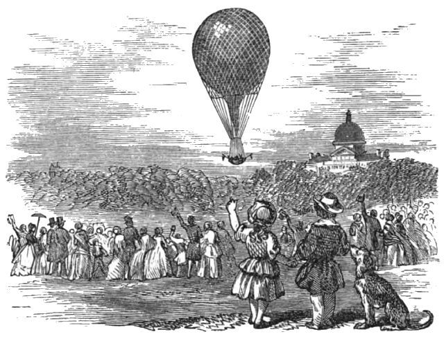 In a field, a group of people are watching a hot air balloon in the sky above