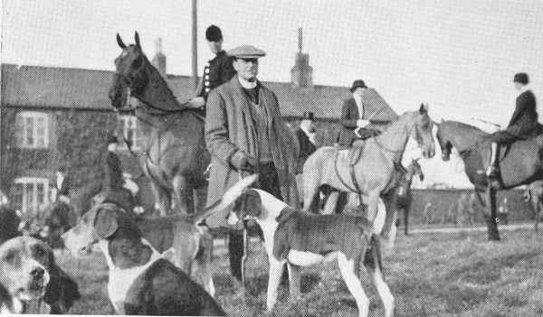 man standing with walking stick, surrounded by hunting dogs with 3 mounted huntsmen behind on horeseback