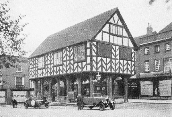 Tudor building with empty open-air market area and second story Tudor design, two 1930’s cars parked in front
