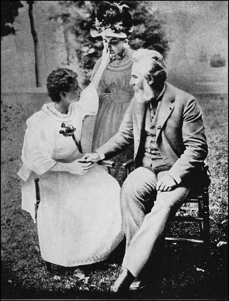 Helen Keller is seated on the left, wearing a white dress. She has her hand at the mouth of Ann Sullivan who is standing behind her. Dr. Bell, seated on the right, has a beard and mustache and is wearing a light colored suit.