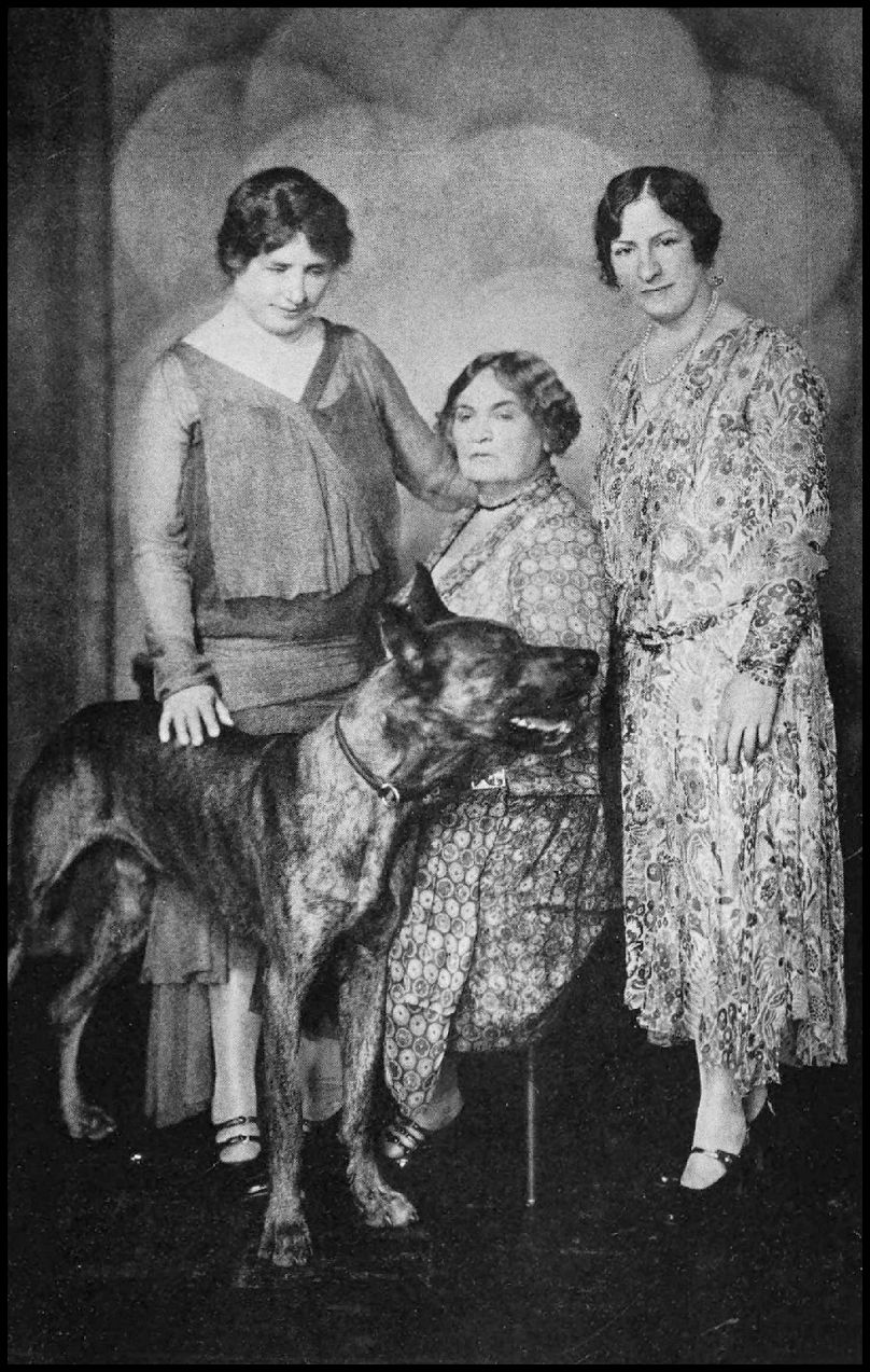 Three women are in the photograph. One woman is seated and the other two are standing on each side of her. The woman in the left has her hand on the back of a large dog.