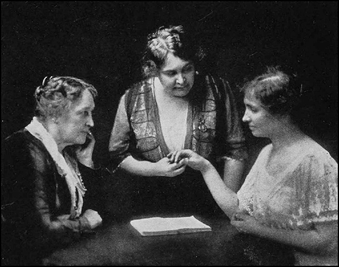 Three women are seated at a table. The woman in the center of is holding the hand of the woman on the right while the woman on the left watches. There is a book laying in the center of the table.
