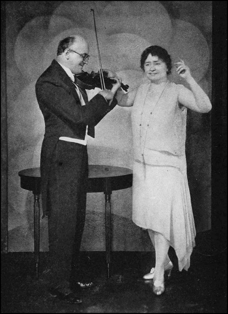 A man, wearing a formal suit, is standing on the left and playing a violin. A woman, wearing a dress, is standing to the right of the man. She has her right hand on the violin and her left hand in the air.