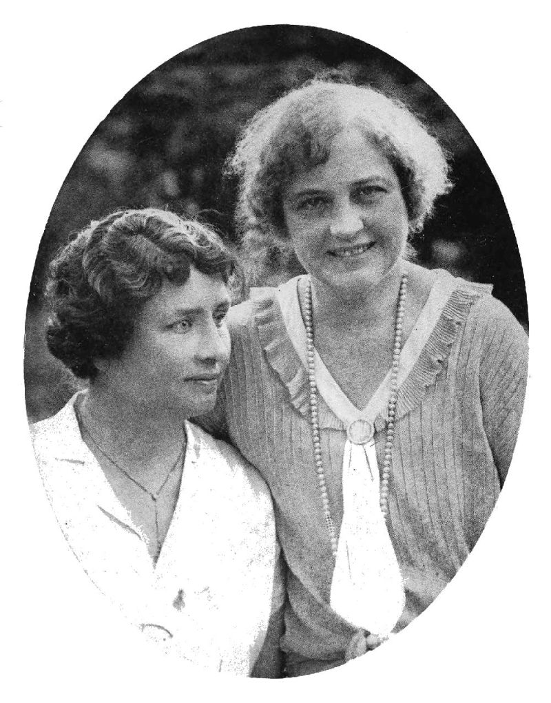 Two women are standing next to each other. The woman on the right is looking into the camera while the woman on the left gazes into the distance.