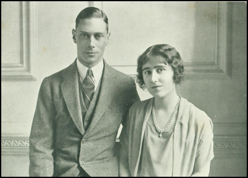 A man wearing a suit is standing next to a woman with short hair wearing a pearl necklace.