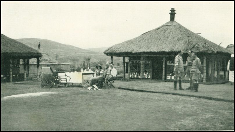 A group of men are standing on the left while another group of people are seated at a table on the right. In the background are thatched roofed huts.