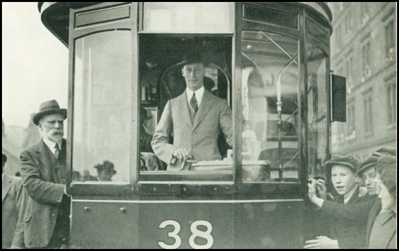 A man is standing inside the engine of a tram and appears to be driving the tram. There are people standing outside on either side of the tram.