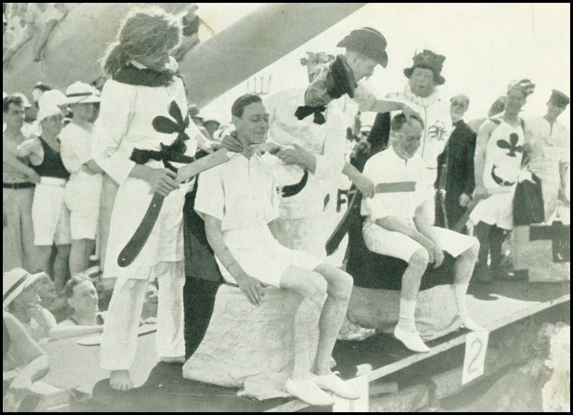 A man is sitting on a stool. Another man is applying shaving cream to the seated man's chin. Another man is holding an oversized shaving razor. There is a crowd of people watching and some people are wearing what appear to be costumes.