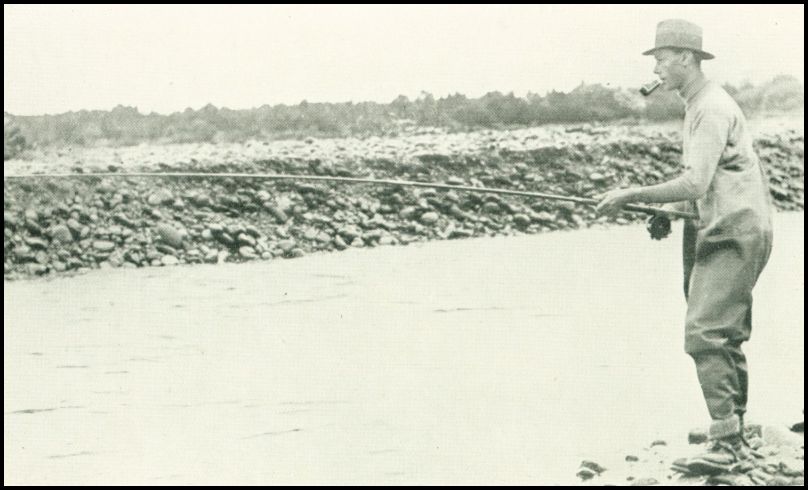 A man, wearing waders, is fly fishing from the bank of a river.