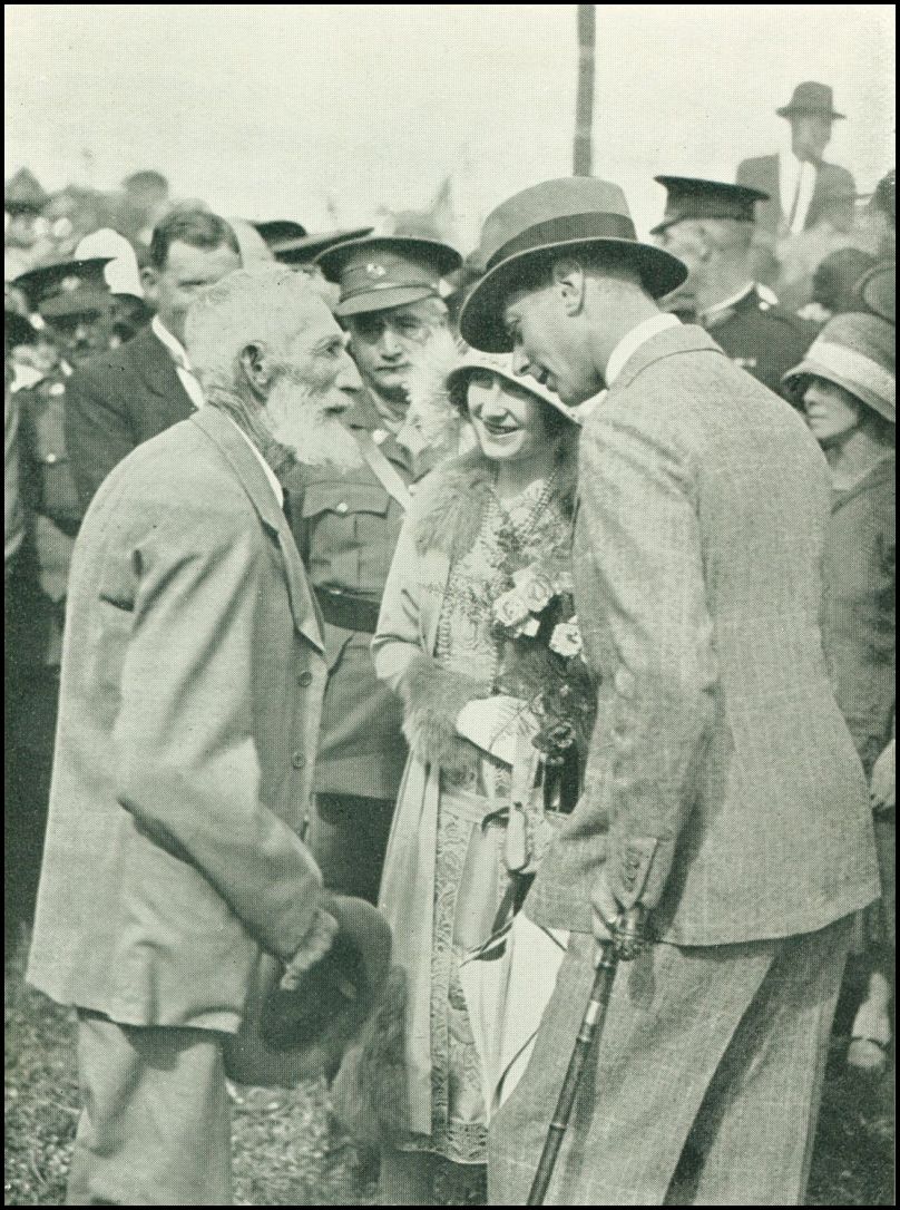 A man wearing a hat is speaking with an older man with a beard. A woman, holding a bouquet of flowers is also in the group. Many people, some in uniform, are in the background.