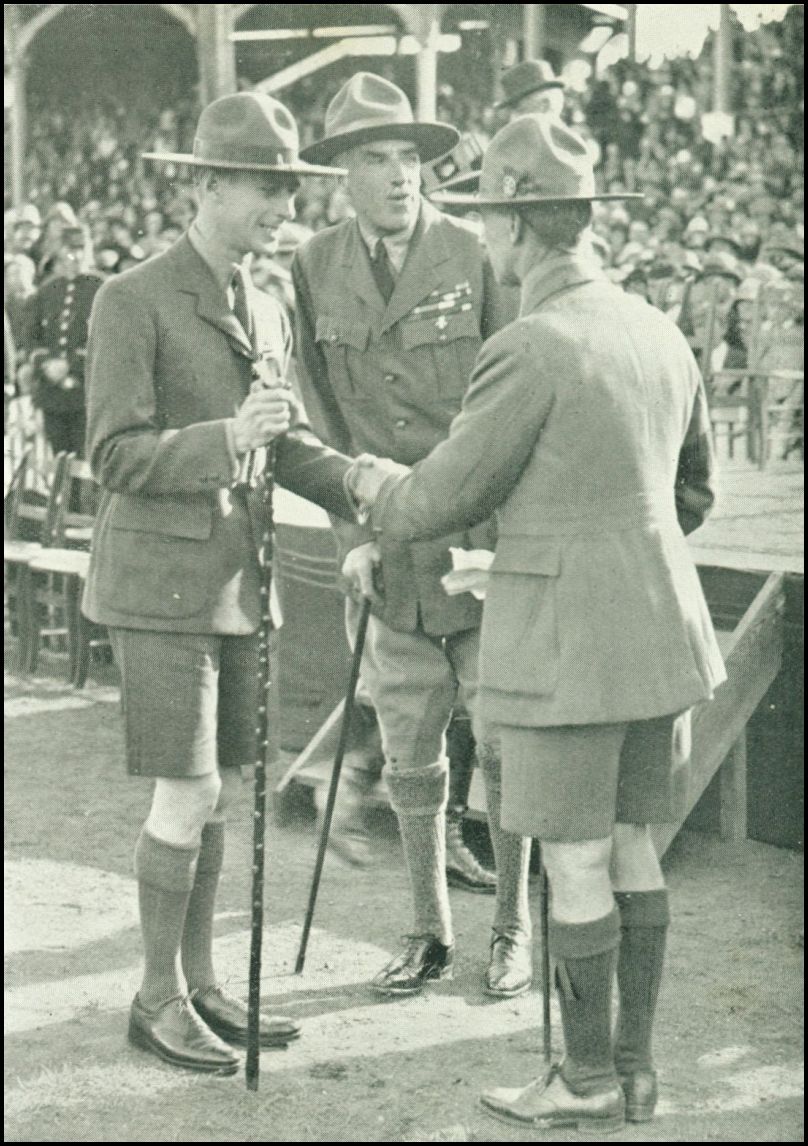 Three men wearing scouting uniforms are standing together. One man is shaking the hand of another man. There is a huge crowd of people in the background.
