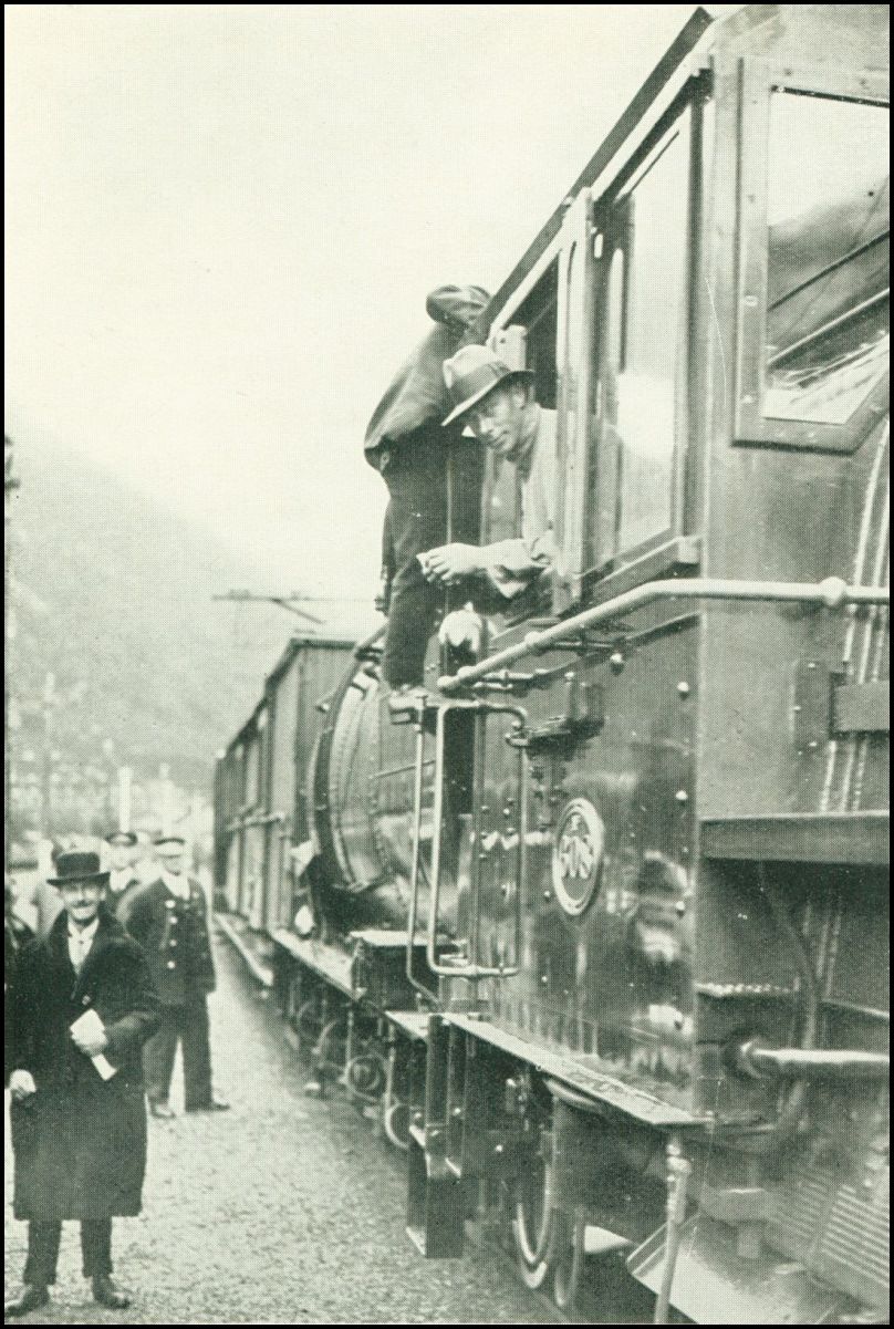 A man is leaning out of the engine window of the train and looking at the camera. There is another man leaning over the top of the engine.
