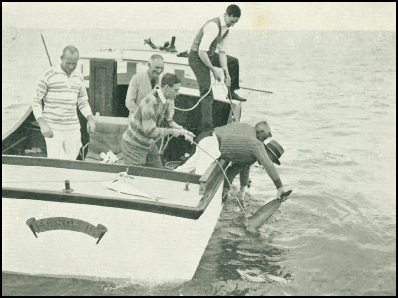 A group of five men are on a fishing boat in the ocean. One man is holding a fishing rod while another man is holding a fish by the tail and attempting to pull it out of the water.
