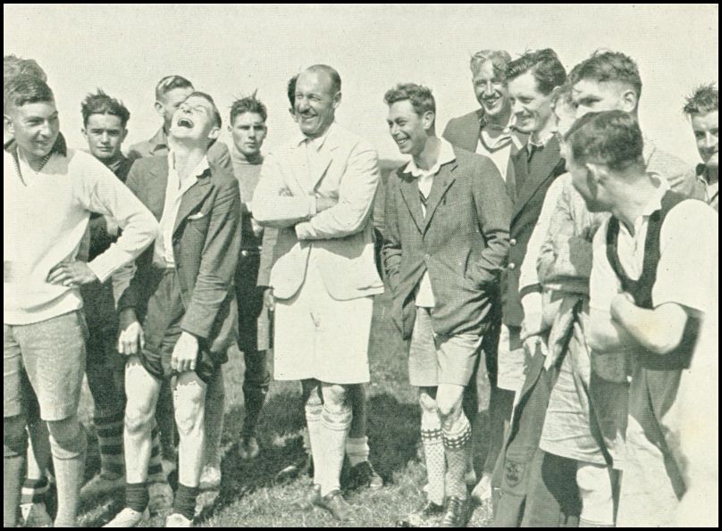 A number of men, all wearing shorts, are standing in a group. All the men are smiling and some of the men are laughing.