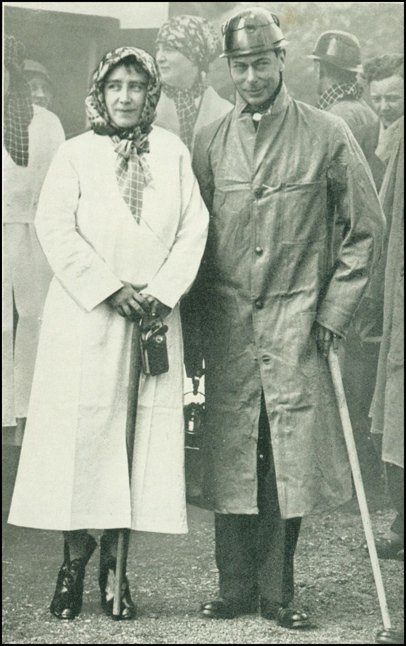 A couple are standing together. The man is wearing a long coat and a hard hat, while the woman is wearing a long coat and a kerchief on her head. There are a number of similarly dressed men and women behind the couple.