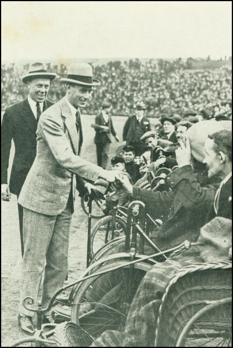 A man wearing a suit and a hat is shaking the hand of other man seated in a wheelchair. There is another man standing behind the first man and a number of other men in wheelchairs. There appears to be a large crowd of people in the background.