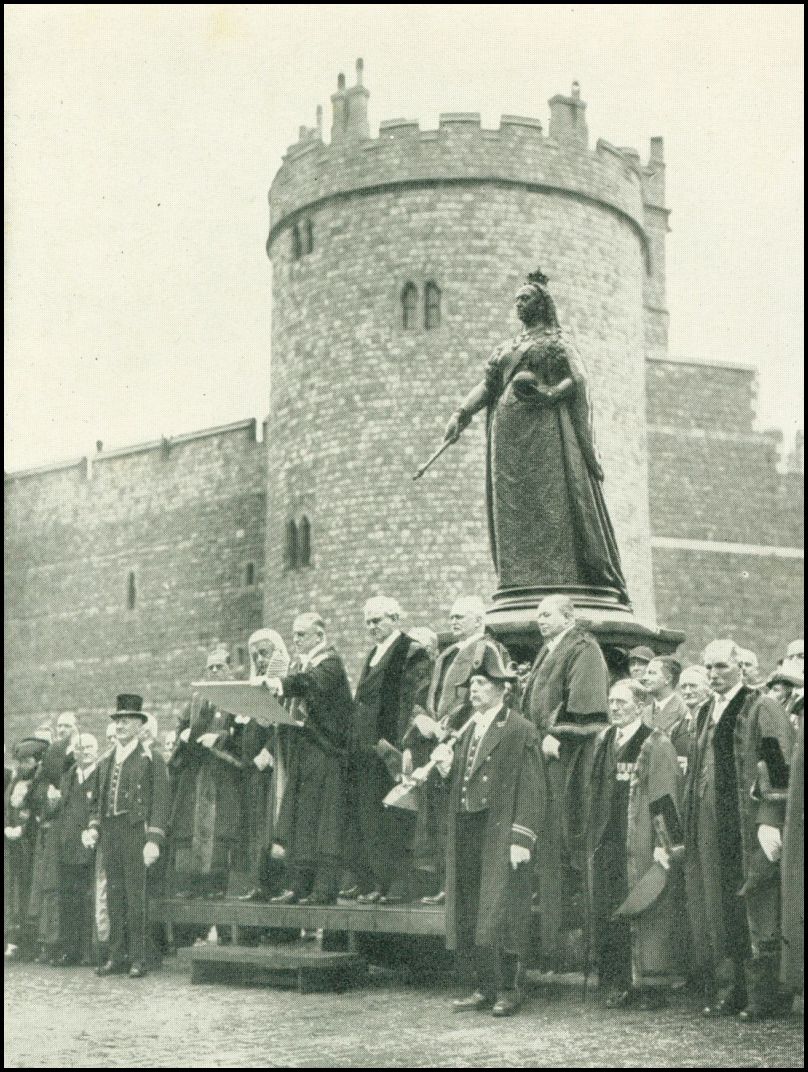 A large group of men, some of whom are on a platform, are standing in front of a statute of a woman wearing a long gown with a crown on her head. There is a castle in the background.