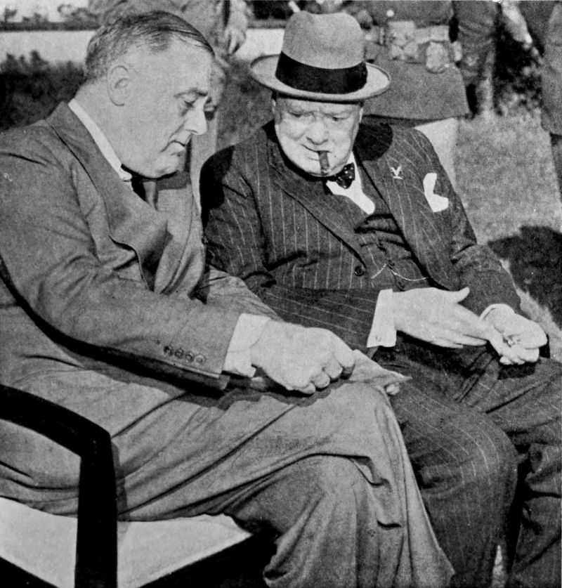 A photo of two men seated on a bench. Both men are wearing suits. The man seated on the right is also wearing a hat and has a cigar in his mouth. The man seated on the left is holding a piece of paper on his knee and both men are looking at the paper.