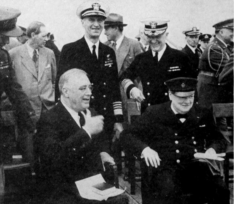 A photo of two men sitting in chairs with many other men standing behind them. The man seated on the right is wearing a uniform with a double row of buttons and a hat. The man seated on the left is wearing a suit and appears to be speaking.
