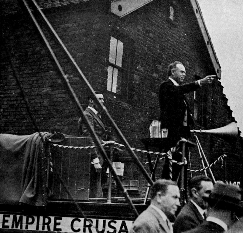 A photo of a man standing on an elevated stage and pointing into the distance. Another man is standing behind him and there is a group of men standing on the bottom right of the photo. Visible on the bottom left are the words-Empire Crusa--.