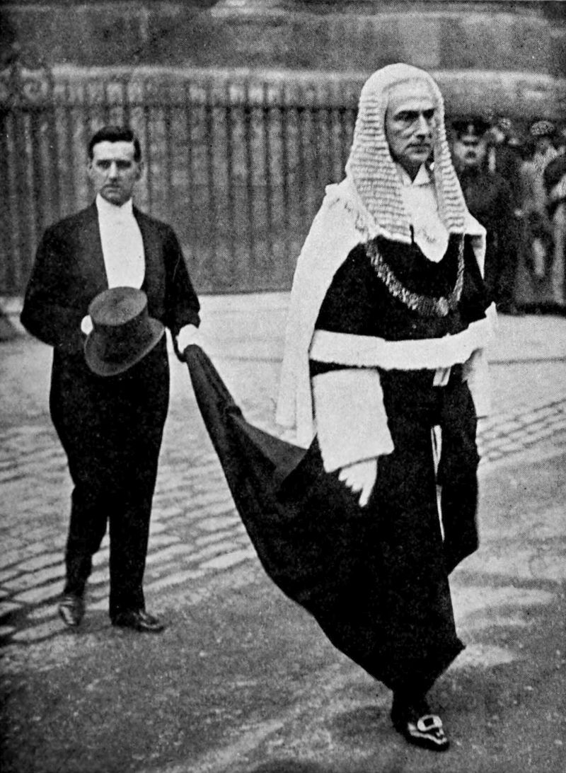 The photo shows two men walking. The man in front is dressed in judical robes and wearing a long wig. The second man is wearing a suit and is holding a top hat in one hand and the robe hem of the man in front in the other hand.