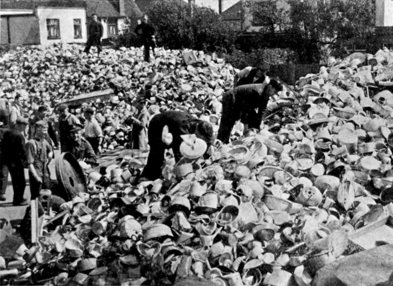 The photo shows a number of people searching through what appears to be piles of scrap metal. There are several buildings in the background.
