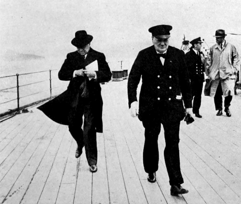 The photo shows two men walking on the deck of a ship which is at sea. The man on the right is wearing a captain's uniform and hat. The man walking behind him is wearing a long coat and hat and is carrying a book. There are three other men in the background on the right.