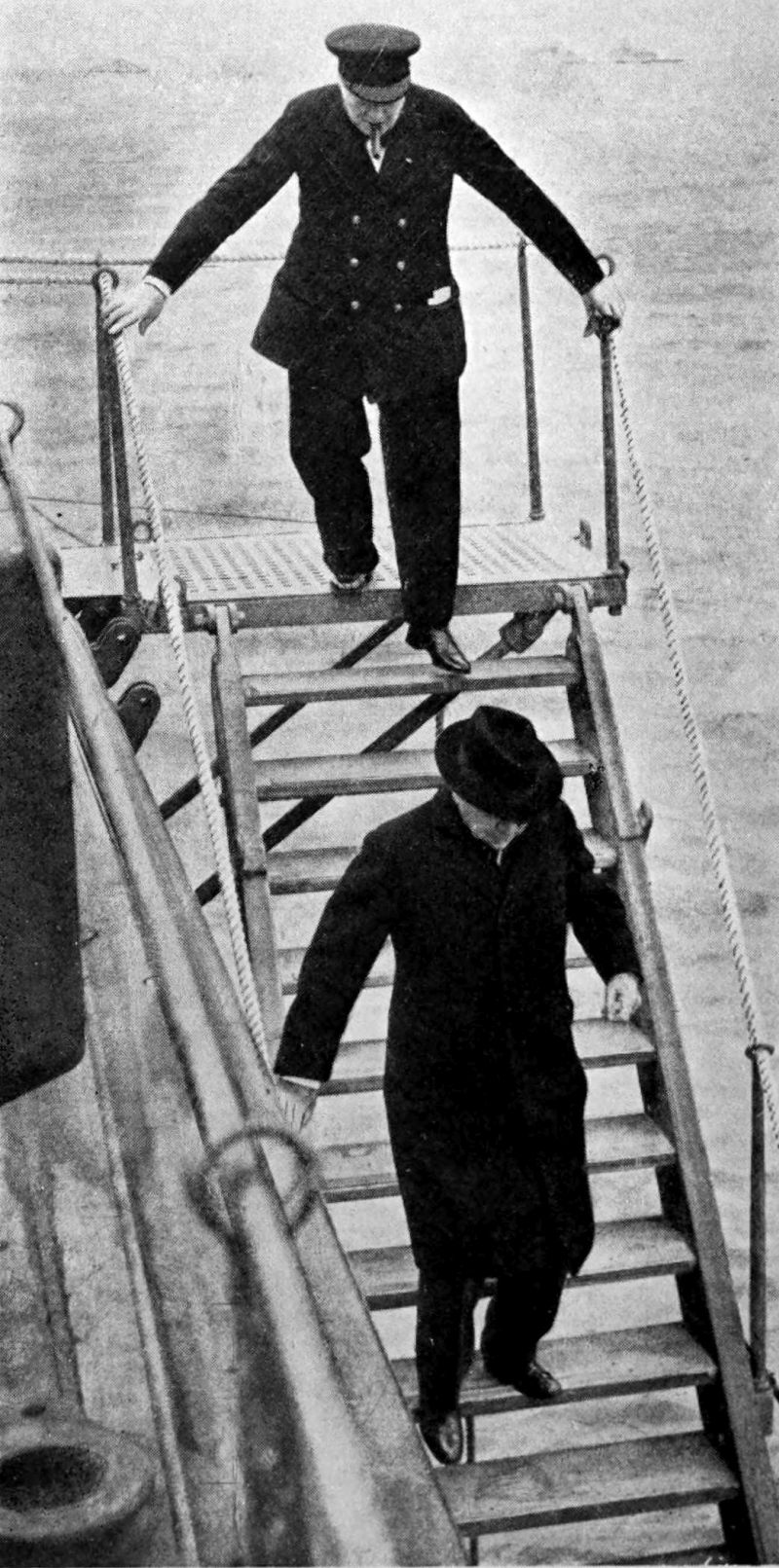 The photo shows two men descending stairs on the deck of a ship at sea. The man on the lower steps is wearing a long coat and a hat. The man at the top of the stairs is wearing a captain's uniform and hat and is smoking a cigar.