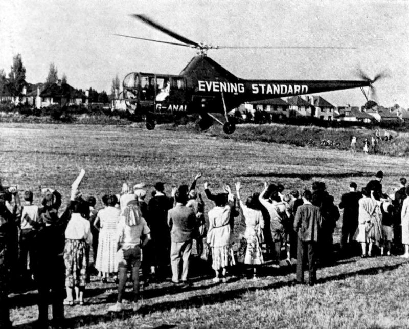 The photo shows a large group of people in the foreground, some of the people are waving. There is a large helicopter with Evening Standard printed on it, landing in the field in the background.