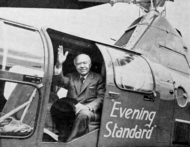 The photo shows a man seated at the open door of a helicopter. The man is waving his hand. Evening Standard is printed on the door of the helicopter.
