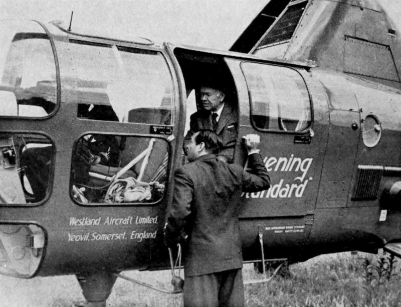 The photo shows the front part of a helicopter. There is a man standing at the open door of the helicopter who appears to be speaking to the people seated inside. Westland Aircraft Limited, Yeovil Somerset, England is printed on the body of the helicopter.