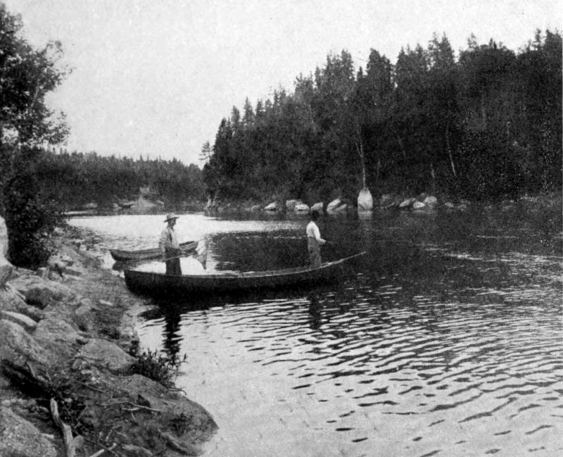 The photo shows two men who appear to be fishing, standing in a canoe on a river. There are tall trees on either bank of the river and rocks lining the sides of the river.