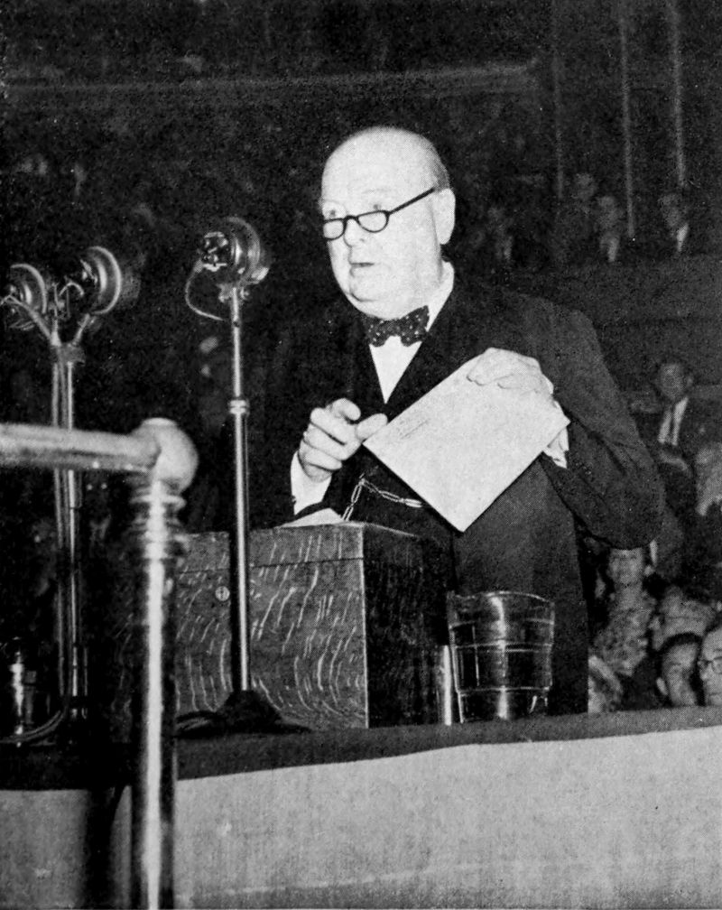 The photo shows a man standing in front of a microphone. The man is wearing glasses and holding a piece of paper in this hand. There is an audience seated behind the man.
