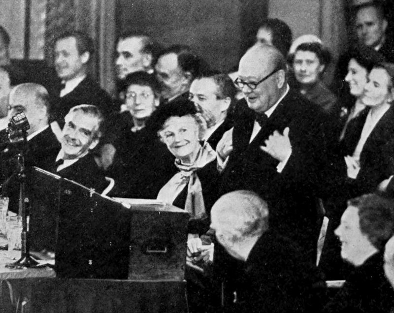 The photo shows a man standing at a lectern. His head is angled downwards as if he is reading something in front of him. There are a number of people seated around him and looking at him. Some of the people are smiling.