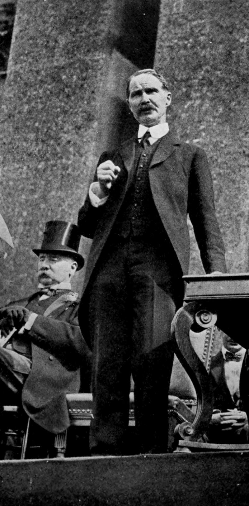 The photo shows a man wearing a suit, standing on a stage. There is another man wearing a top hat seated to the left of the standing man.