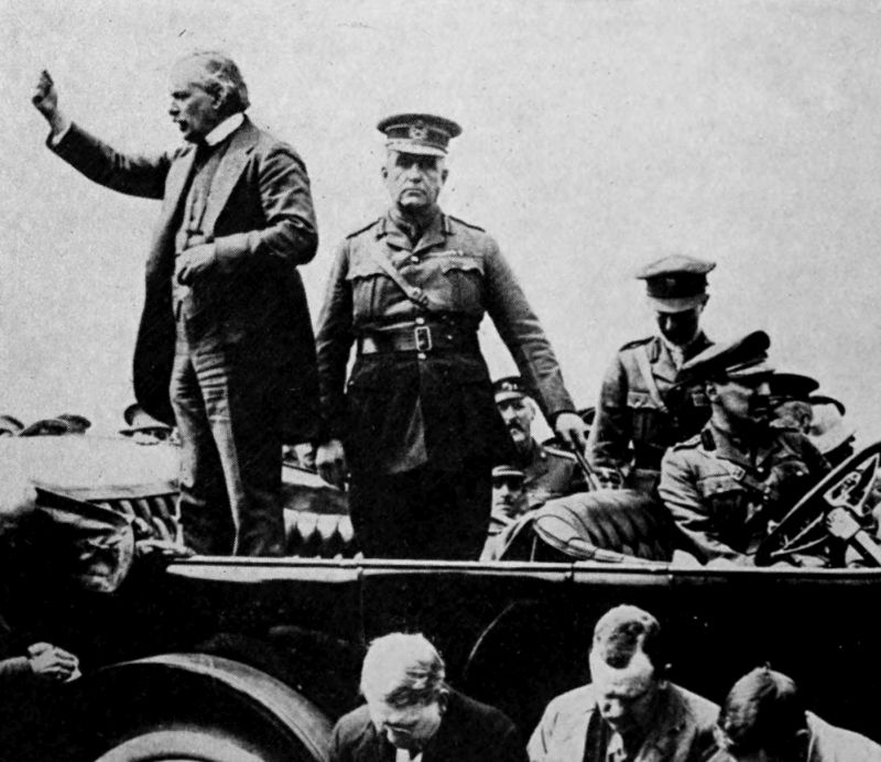 The photo shows three men in a open topped vehicle. The man on the left appears to be waving his right hand. The man in the middle is wearing a uniform and looking into the distance. The man on the right is seated and looking to the right. There are three other people with bent heads in the foreground and several other people in the background.