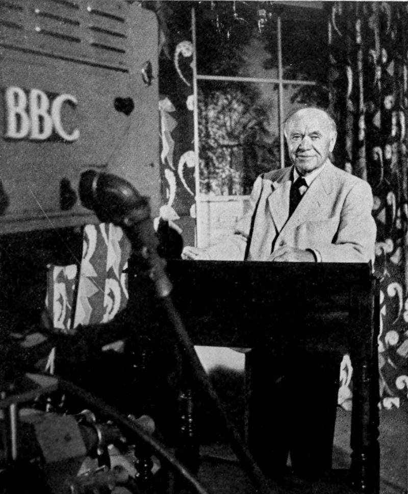 The photo shows a man wearing a light colored suit standing at a lectern. There is a camera with BBC written on it facing the man.