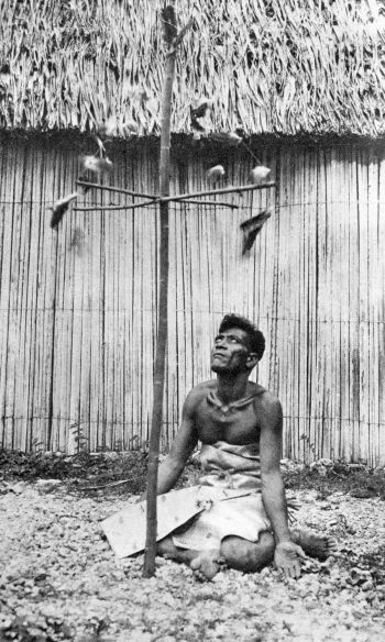 man sitting wearing tapa-cloth waistcloth palms open looking up a pole with fluttering carved objects attached