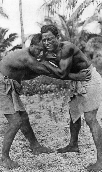 two men barefoot and wearing waistcloths wrestling on coral sand
