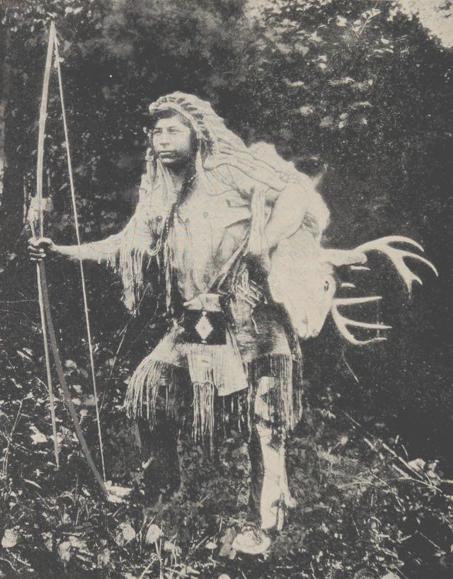 Photo of a Native American brave dressed in full regalia and holding a long bow. He has a deer head with antlers draped over his shoulders.