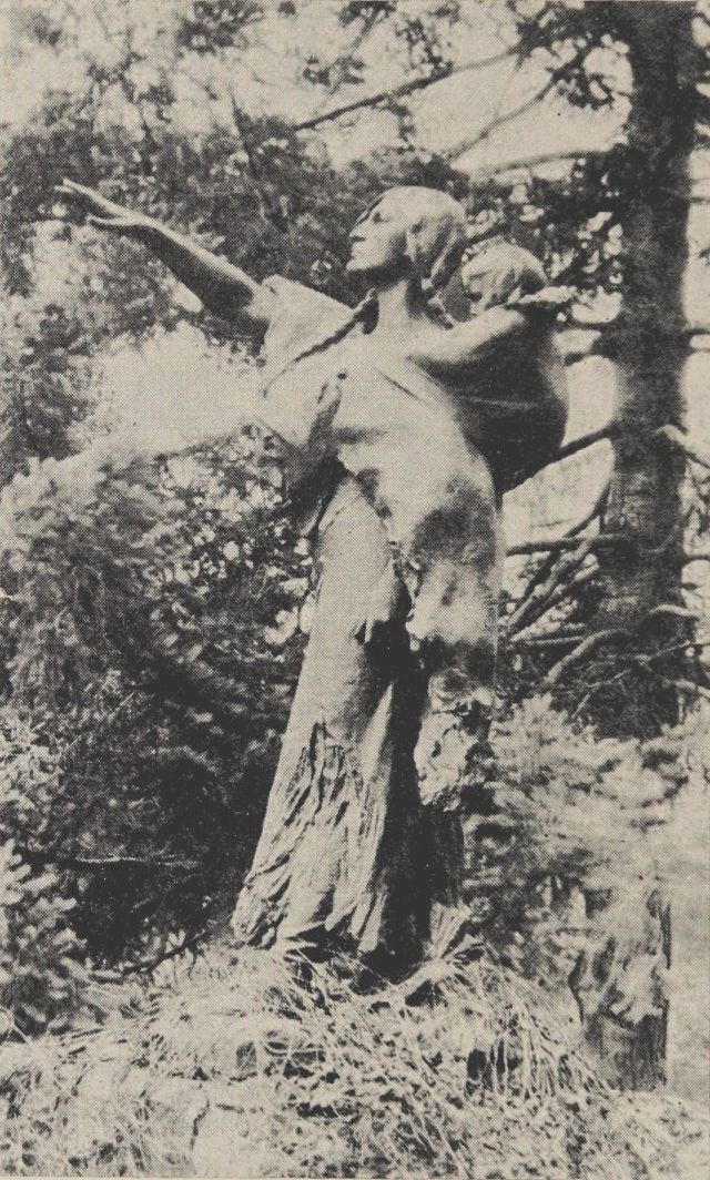 Photo of a Native American woman in a forest setting, carrying a child on her back and pointing her arm into the distance.