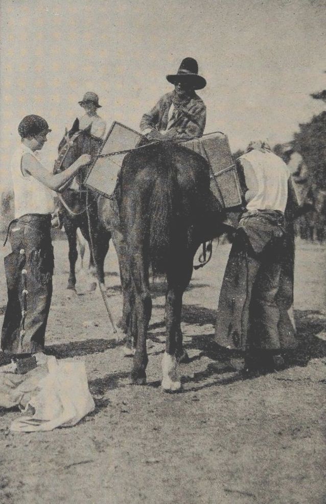 Photo of two men loading a packhorse while another man, seated on a horse watches. A fourth man, in the background, is also seated on a horse.