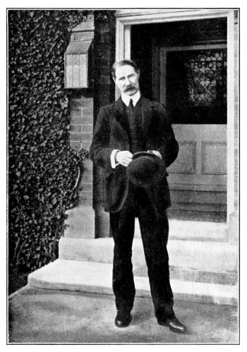 man with moustache in 3-piece suit standing in a doorway holding his hat
