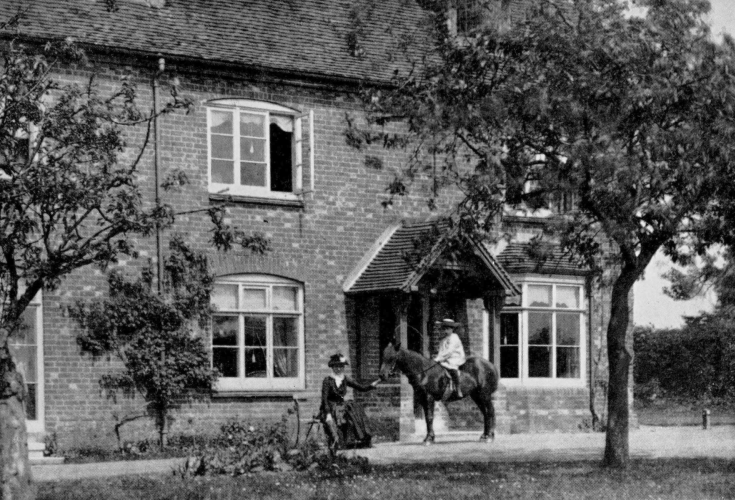 young boy wearing a hat sitting on a horse that seated mother is holding in front of brick two story house