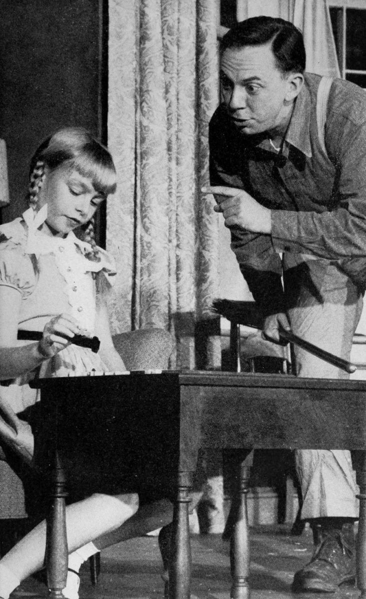 Rhoda seated at a small table with a man leaning over her pointing a finger at her