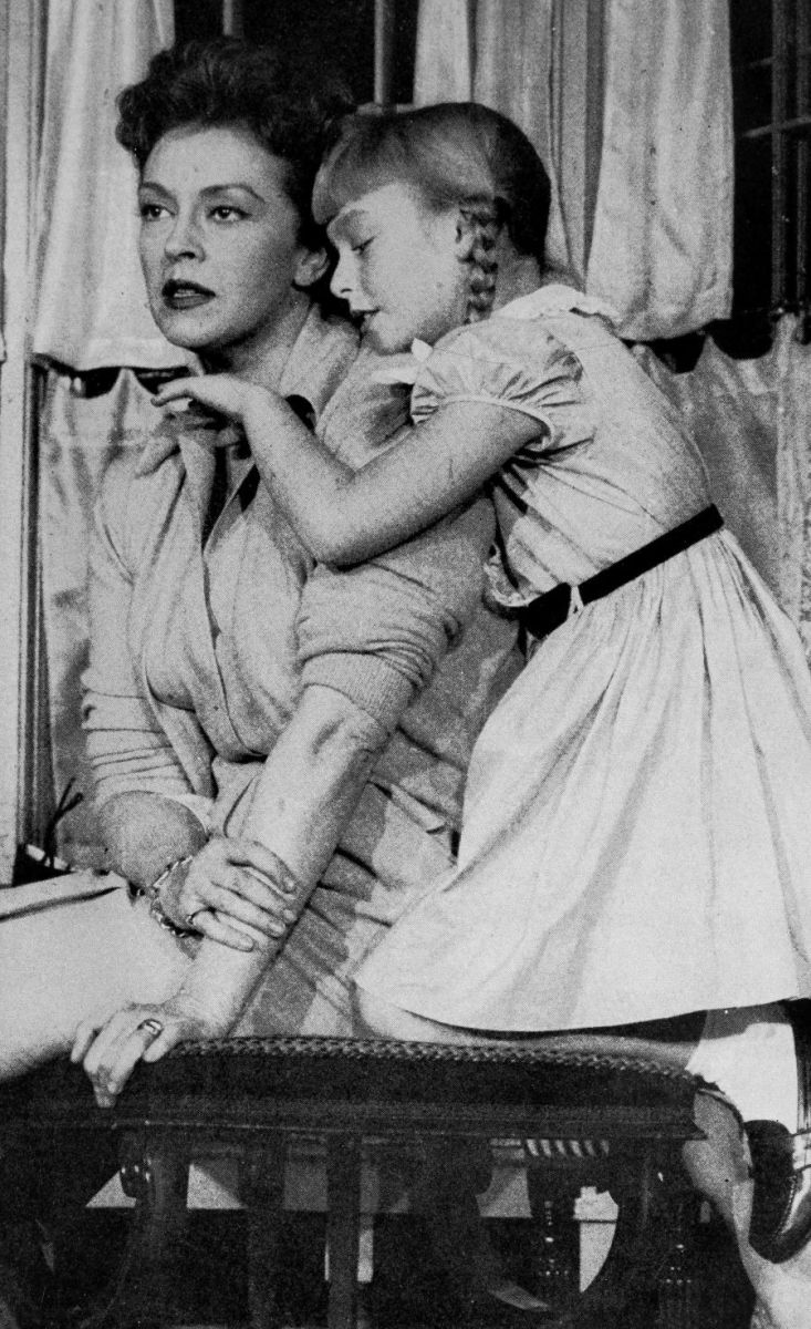 Christine seated and staring straight ahead into the distance while Rhoda who is standing, hugs and fawns over her