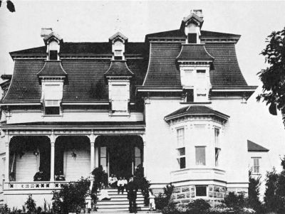 2 story wood house with gables with man standing at front steps and other sitting on porch